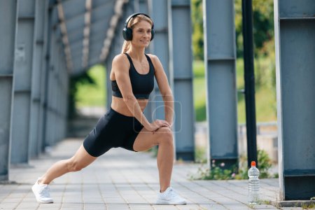 Téléchargez les photos : Femme de fitness souriante portant des écouteurs travaillant et étirant les jambes en plein air. - en image libre de droit
