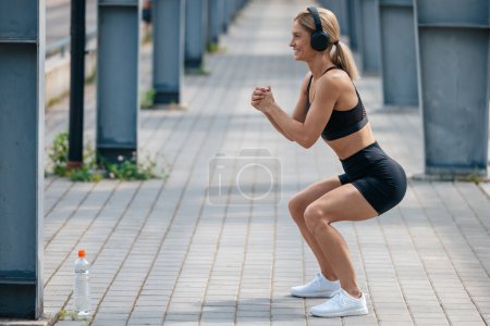 Foto de Mujer en forma deportiva con auriculares escuchando música en cuclillas al aire libre haciendo ejercicios de fitness en la mañana de verano. - Imagen libre de derechos