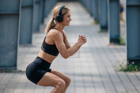 Photo for Athletic woman with headphones listening music squatting outdoor doing fitness exercises in summer morning. - Royalty Free Image