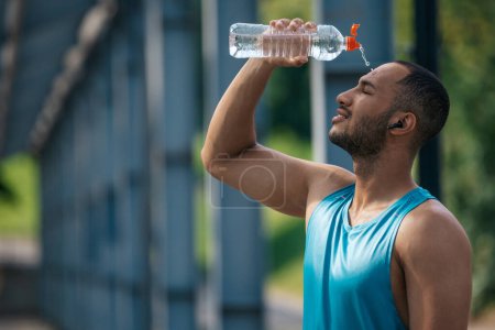Foto de Después del entrenamiento. Joven atleta de piel oscura vertiendo un poco de agua refrescante sobre sí mismo después del entrenamiento - Imagen libre de derechos