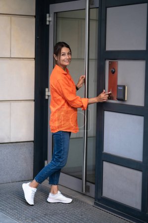 Téléchargez les photos : Femme brune portant une chemise orange et un jean composer un interphone pour entrer dans un bâtiment. - en image libre de droit