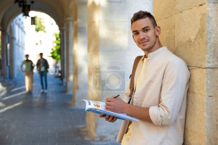Téléchargez les photos : Étudiant. Beau jeune homme debout près du mur avec un manuel dans les mains - en image libre de droit