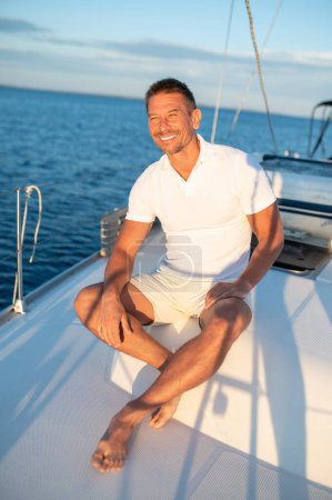 Téléchargez les photos : Sur un yacht. Homme en t-shirt blanc assis sur un yacht avec une corde dans les mains - en image libre de droit