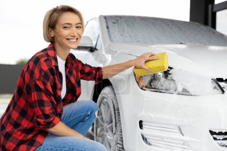 Foto de Lavando un auto. Mujer rubia con camisa a cuadros lavando un coche blanco - Imagen libre de derechos