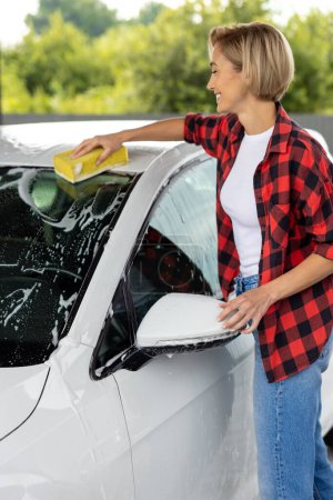 Téléchargez les photos : Lavage de voiture. Blonde jeune femme lavage pare-brise de voiture et regarder impliqué - en image libre de droit