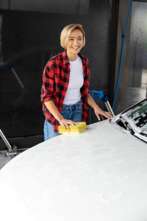 Téléchargez les photos : Lavage de voiture. Femme en chemise rouge nettoyage du capot de voiture - en image libre de droit