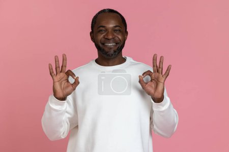 Foto de Hombre negro feliz vistiendo sudadera blanca mostrando signo bien aislado sobre fondo rosa. - Imagen libre de derechos