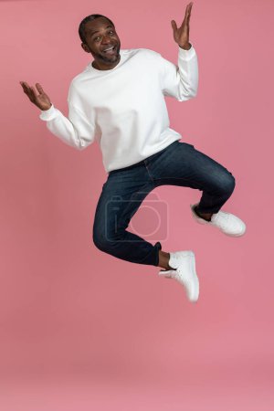 Foto de Hombre negro lleno de alegría vistiendo sudadera blanca saltando alto aislado sobre fondo rosa. - Imagen libre de derechos