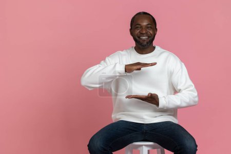 Téléchargez les photos : Homme noir joyeux portant sweat blanc montrant l'espace vide avec les mains grande taille isolé sur fond rose. - en image libre de droit