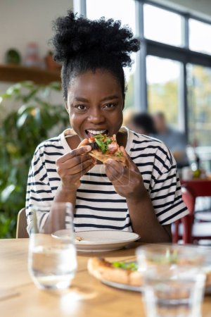 Téléchargez les photos : Mignonne jeune femme à la peau foncée dégustant une pizza - en image libre de droit