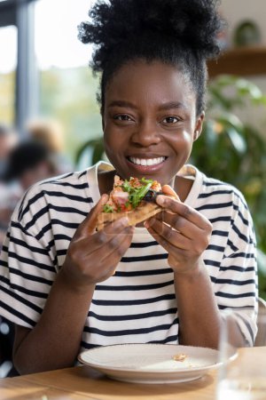 Téléchargez les photos : Mignonne jeune femme à la peau foncée dégustant une pizza - en image libre de droit