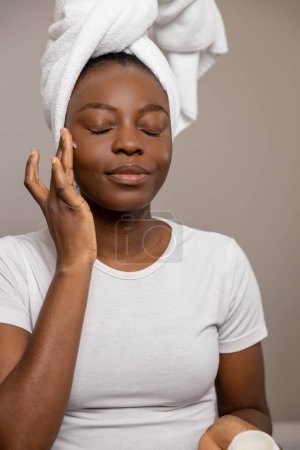 Foto de Mujer satisfecha aplicando crema de cuidado diario cerrando los ojos de pie con toalla en la cabeza contra la pared marrón - Imagen libre de derechos