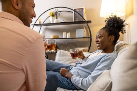 Téléchargez les photos : Couple heureux parlant boire du thé à partir de verres assis sur le canapé dans le salon - en image libre de droit