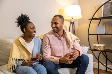 Téléchargez les photos : Un bel homme et une femme souriante assis sur le canapé à la maison et buvant du thé à partir de lunettes - en image libre de droit