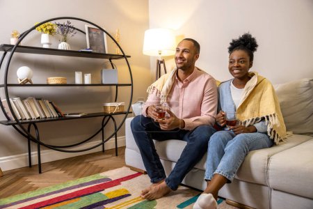 Téléchargez les photos : Un bel homme et une femme souriante assis sur le canapé à la maison et buvant du thé à partir de lunettes - en image libre de droit