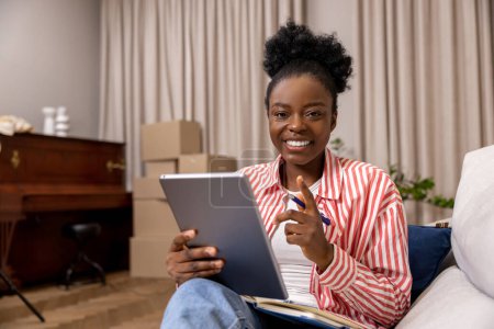Foto de Mujer feliz positiva leyendo notas de libro electrónico mientras está sentada en casa después de mudarse. - Imagen libre de derechos