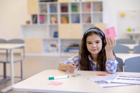 Téléchargez les photos : Petite élève assise au bureau dans la salle de classe de l'école faisant des tâches, montrant des notes en papier. - en image libre de droit