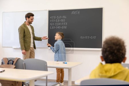 Téléchargez les photos : Petit garçon élève debout dans la salle de classe tout en apprenant les mathématiques leçon avec son professeur. - en image libre de droit