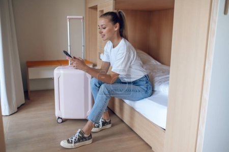 Téléchargez les photos : Voyageur femme utilisant téléphone portable tout en se détendre sur le lit dans la chambre d'hôtel. - en image libre de droit