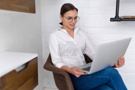 Photo for Young confident woman working on laptop in armchair indoors. - Royalty Free Image