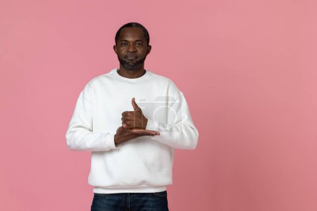 Foto de Hombre negro mudo sordo con sudadera blanca haciendo gestos de lenguaje de señas aislado sobre fondo rosa. - Imagen libre de derechos