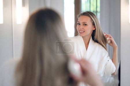 Foto de Haciendo pelo. Mujer bonita de pelo largo haciendo pelo en el espejo - Imagen libre de derechos