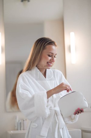 Photo for Morning beauty. Smiling young woman in white robe holding a bag cosmetics - Royalty Free Image