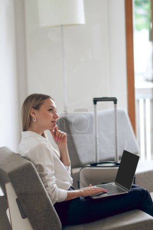 Téléchargez les photos : Femme attrayante avec valise et ordinateur portable assis sur un fauteuil dans son appartement. - en image libre de droit
