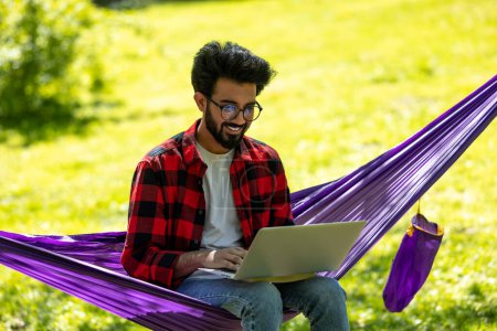 Foto de Hombre con camisa a cuadros sentado en hamaca y trabajando en el ordenador portátil - Imagen libre de derechos
