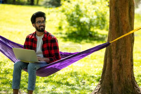 Téléchargez les photos : Homme en chemise à carreaux assis dans un hamac et travaillant sur un ordinateur portable - en image libre de droit