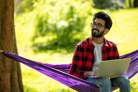 Téléchargez les photos : Satisfait jeune homme hindou barbu avec ordinateur portable dans le parc - en image libre de droit