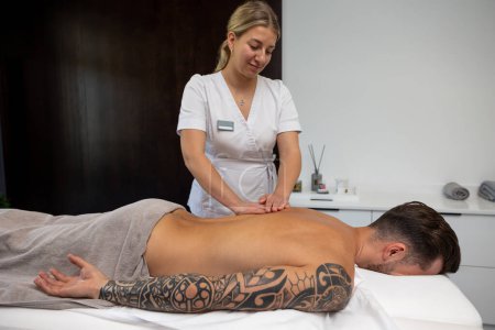 Massage du dos. Jeune homme avec un gros tatouage ayant séance de massage du dos dans un salon
