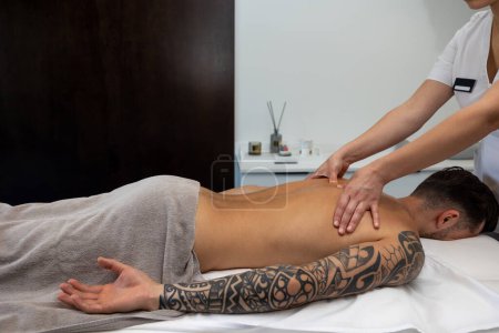 Foto de Masaje de espalda. Joven hombre con un gran tatuaje teniendo sesión de masaje de espalda en un salón - Imagen libre de derechos