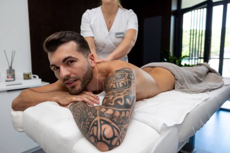 Téléchargez les photos : Massage du dos. Jeune homme avec un gros tatouage ayant séance de massage du dos dans un salon - en image libre de droit