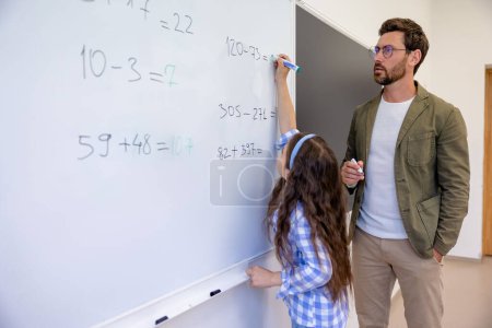 Photo for Primary school teacher watching pupil writing on white board in classroom. - Royalty Free Image