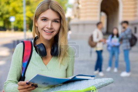 Téléchargez les photos : Étudiante blonde avec des livres se préparant pour les classes posant en plein air près du collège. - en image libre de droit