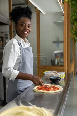 Téléchargez les photos : Femme à la peau foncée souriante préparant une pizza et ayant l'air satisfaite - en image libre de droit