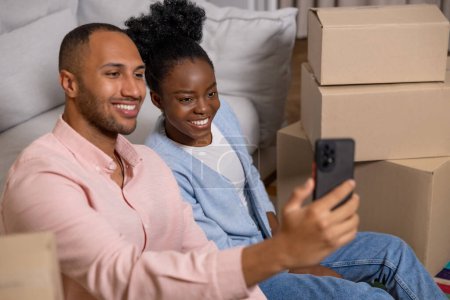 Photo for Adult couple taking selfie with smartphone in their new home after relocation surrounded with carton boxes - Royalty Free Image