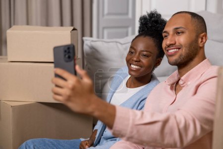 Photo for Cheerful man and woman taking selfie with smartphone in their new home after relocation surrounded with carton boxes - Royalty Free Image