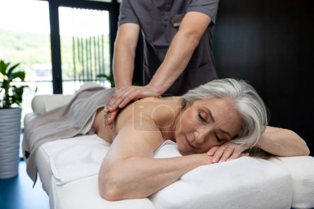 Téléchargez les photos : Massage du dos. Femme mûre aux cheveux gris allongée sur un canapé et ayant une séance de massage du dos - en image libre de droit