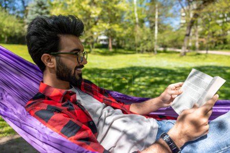 Téléchargez les photos : Satisfait jeune homme dans l'hamac lisant un livre - en image libre de droit