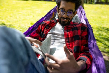 Téléchargez les photos : Jeune homme aux cheveux foncés lisant un livre tout en restant allongé dans un hamac - en image libre de droit