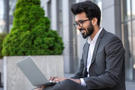 Téléchargez les photos : Jeune homme d'affaires hindou travaillant sur un ordinateur portable assis près de l'immeuble de bureaux - en image libre de droit
