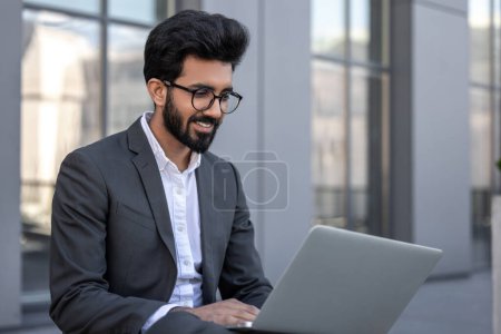 Photo for Young hindu businessman working on a laptop while sitting near the office building - Royalty Free Image