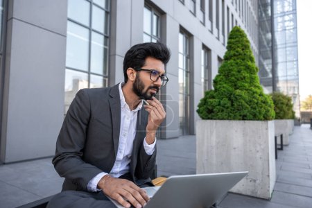 Photo for Young hindu businessman working on a laptop while sitting near the office building - Royalty Free Image