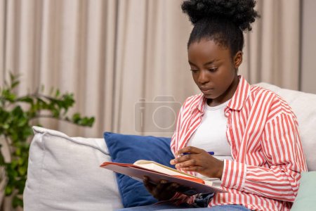 Photo for Adult serious concentrated woman writing her personal notes into her diary while sitting on sofa in living room. - Royalty Free Image