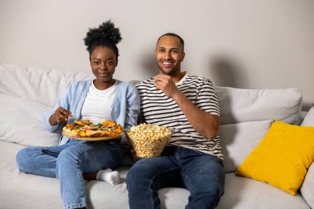 Foto de Alegre pareja comiendo pizza y palomitas de maíz viendo comedia película sentado en sofá interior - Imagen libre de derechos