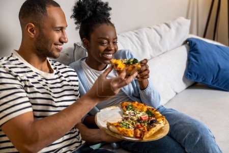 Téléchargez les photos : Happy couple manger une collation de pizza à la maison et rire tout en se reposant sur le canapé - en image libre de droit