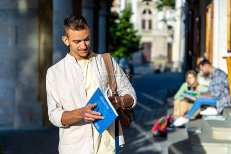 Téléchargez les photos : Attrayant étudiant universitaire debout avec livre ayant une pause entre les classes. - en image libre de droit