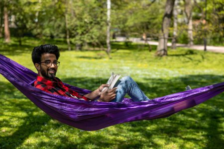 Joven hombre hindú en camisa a cuadros se siente relajado en hamaca en el parque